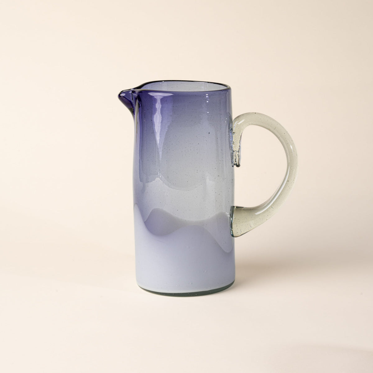Violet and clear handblown glass pitcher with organic wave design with clear handle, casting a soft golden shadow on a beige backdrop.